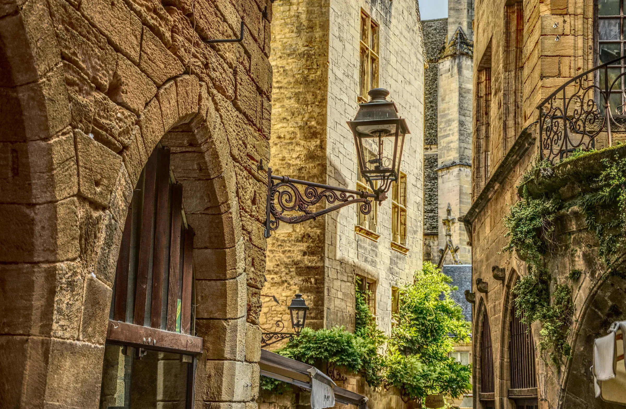 A sunlit medieval street in Sarlat-la-Caneda, honey-coloured limestone houses with an ornate wrought-iron street lantern in the foreground and the Gothic cathedral bell tower rising behind, greenery spilling from a balcony