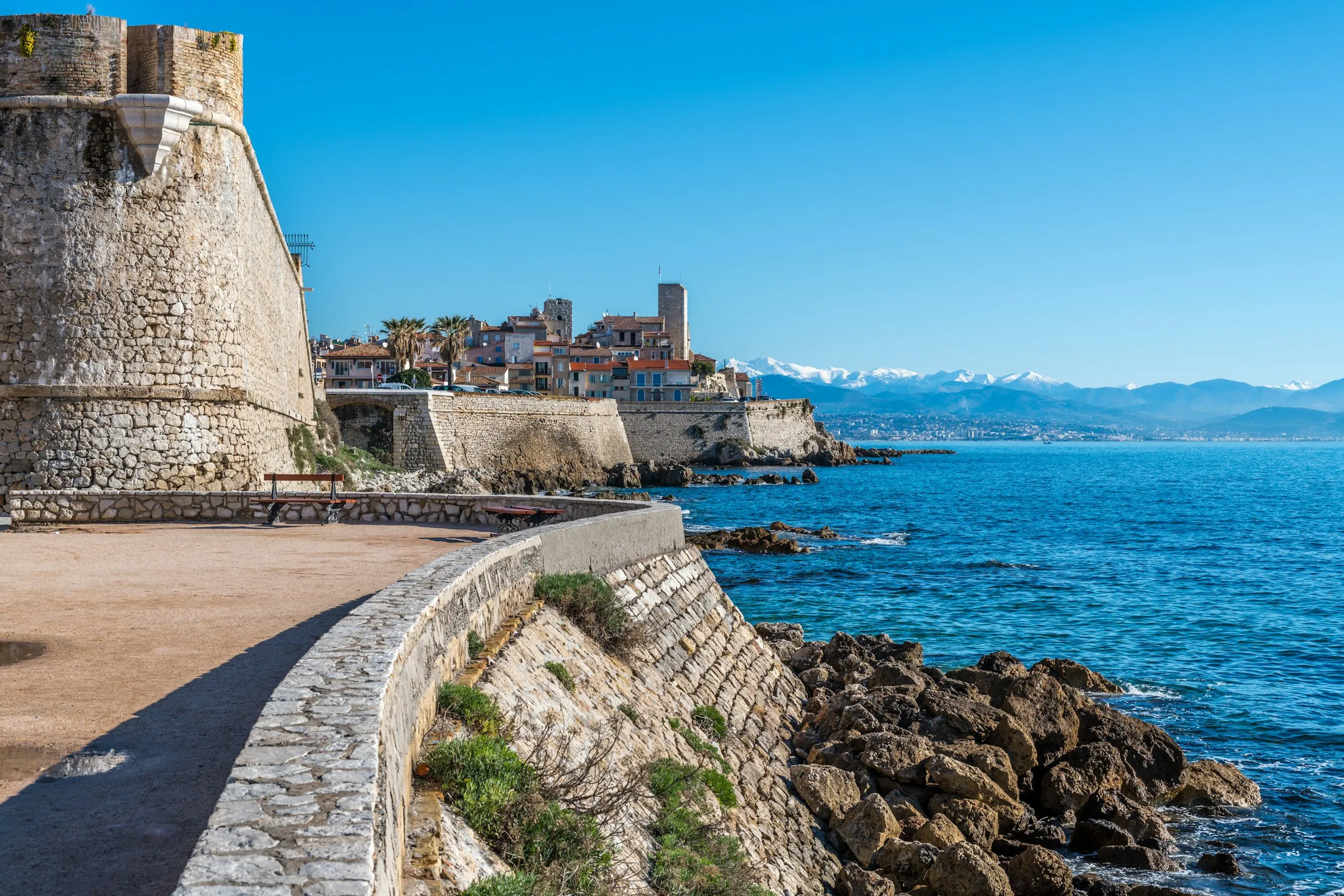 Sunlit seafront rampart promenade curving past Antibes' honey-stone walled old town, with the bastion, terracotta rooftops and a distant snow-capped Alpine range beyond the deep-blue Mediterranean.