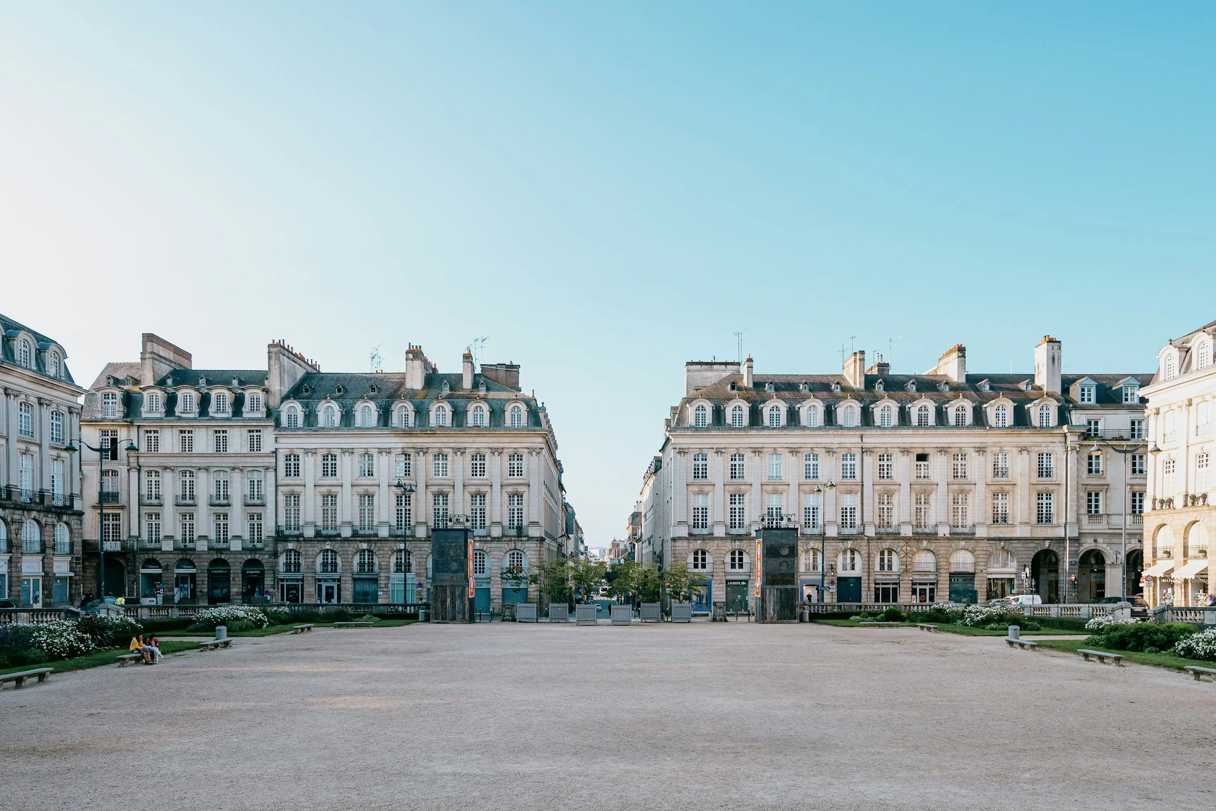 A grand symmetrical pair of 18th-century cream-stone buildings with tall grey slate mansard roofs and arcaded shopfronts framing a perspective street, seen across a wide open gravel esplanade under a clear blue sky in central Rennes
