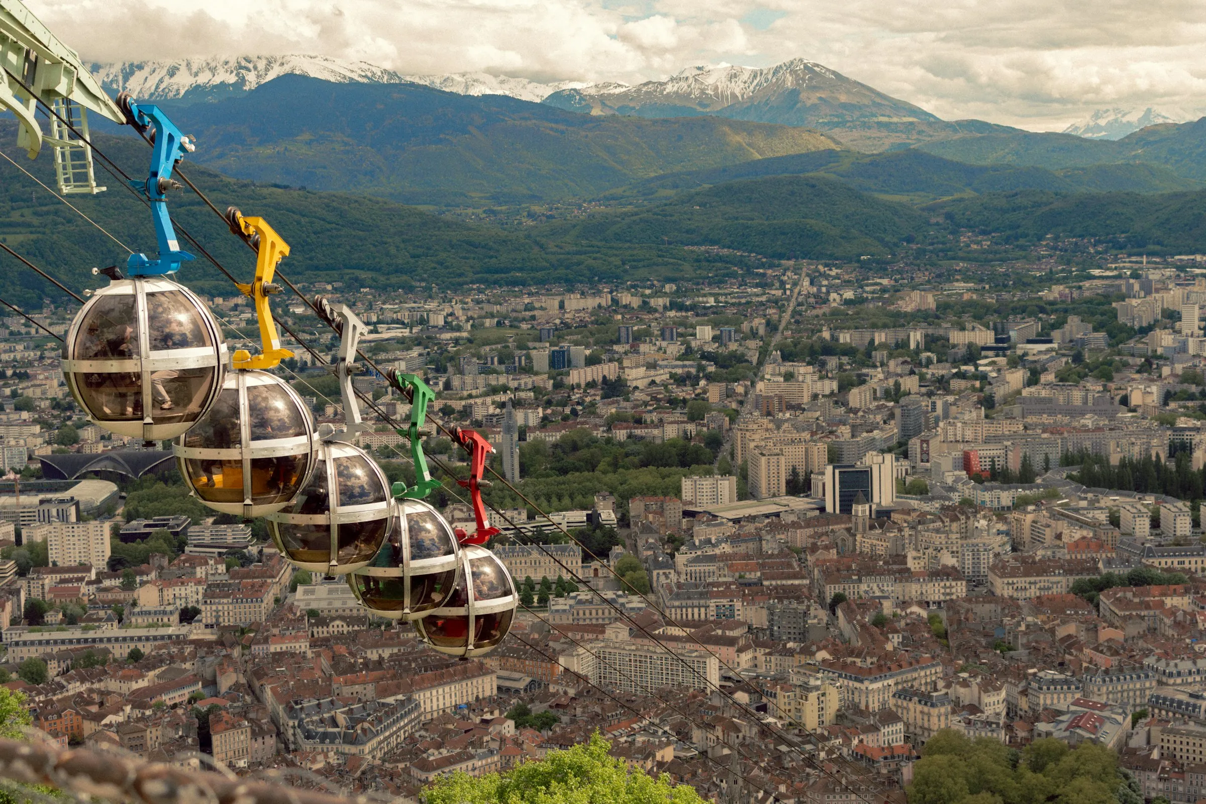 Grenoble's signature 'Bulles' cable car — a line of colored spherical cabins in blue, yellow, green and red — climbing its cable high above the city's dense rooftops, with the snow-dusted Belledonne Alps filling the horizon under a bright clouded sky.