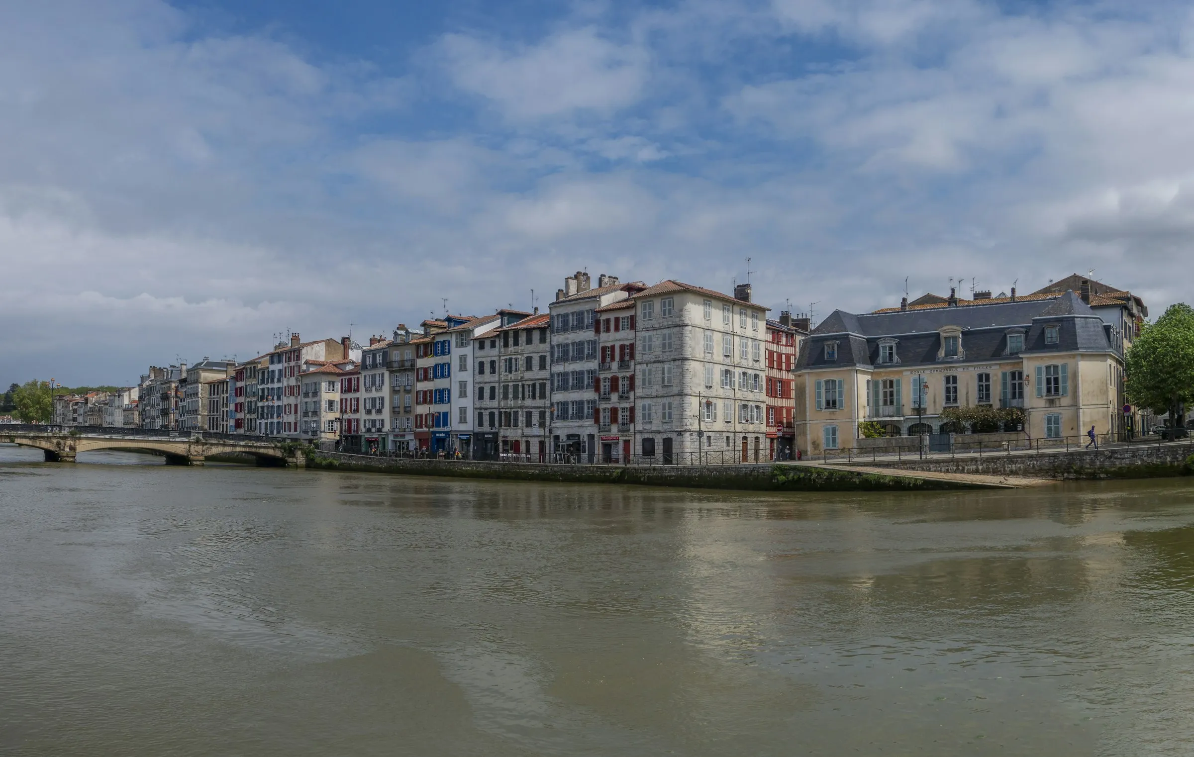 A bright, sunlit row of tall colourful Basque townhouses with red and blue shutters lining the Nive riverfront in Bayonne, a stone bridge to the left and the old girls' school building at the water's edge under a blue sky.