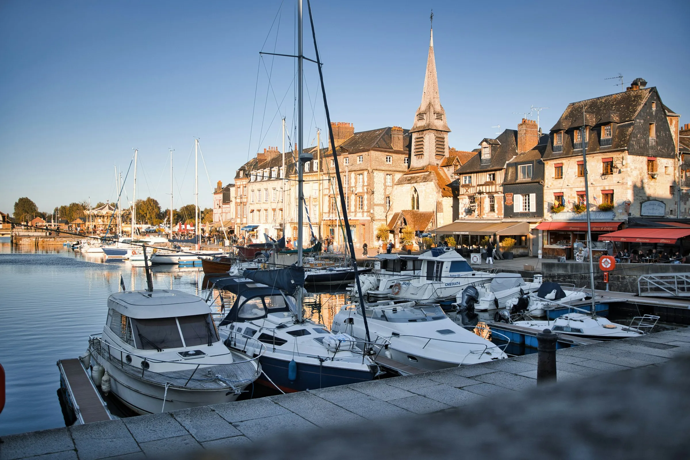 Honfleur's Vieux Bassin at golden hour, with sailboats and pleasure boats moored along the quay below the slate-spired Saint-Etienne church and tall stone quayside houses, warm evening light reflecting on the water