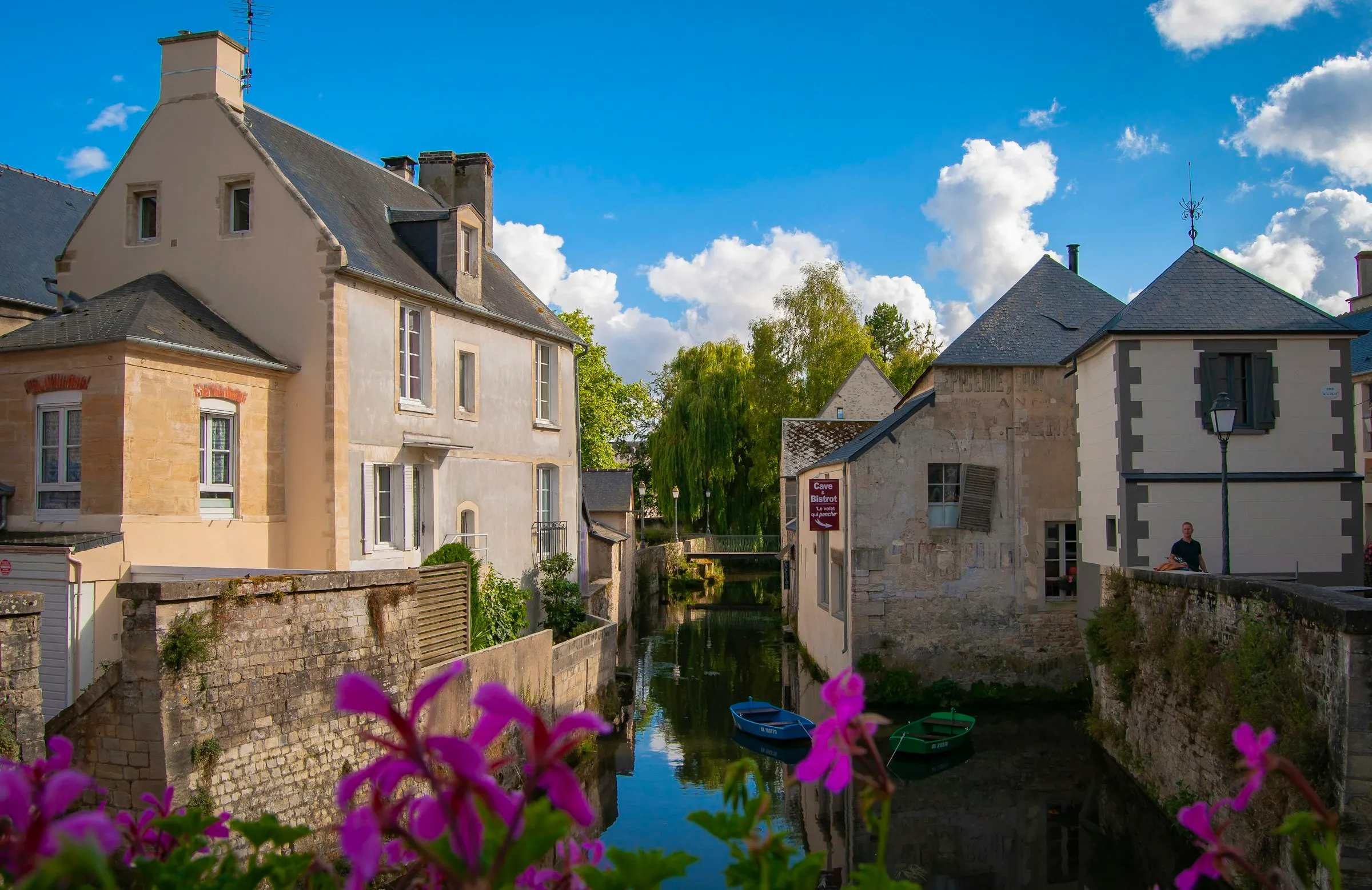 The Aure river winding between Bayeux's old stone houses, two small wooden rowboats moored on the calm water, bright purple geraniums in the foreground and a blue sky scattered with white clouds overhead.