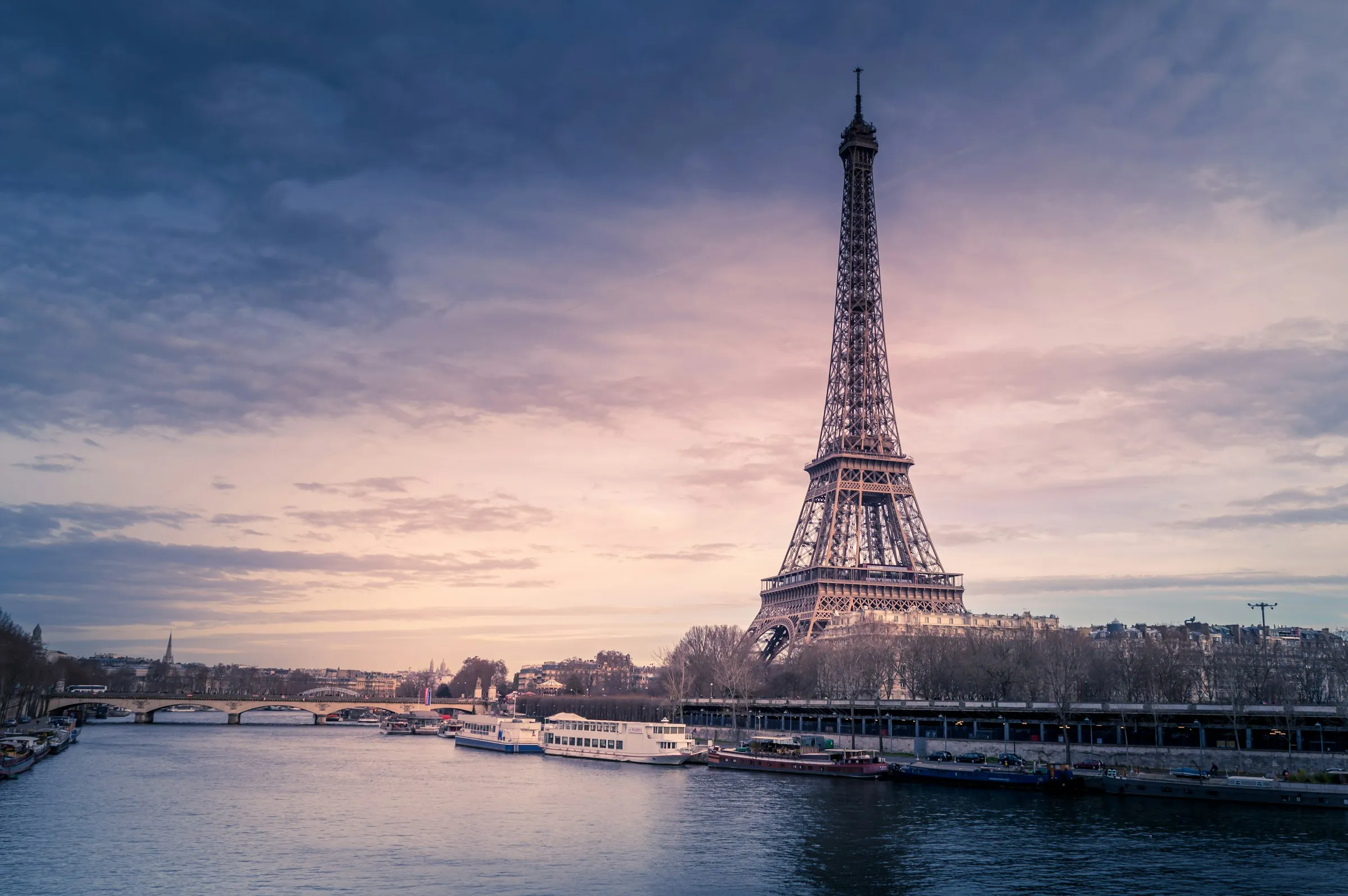The Eiffel Tower rising above the Seine at dusk, with sightseeing boats moored along the quay and a bridge crossing the river under a pink-and-gold sky
