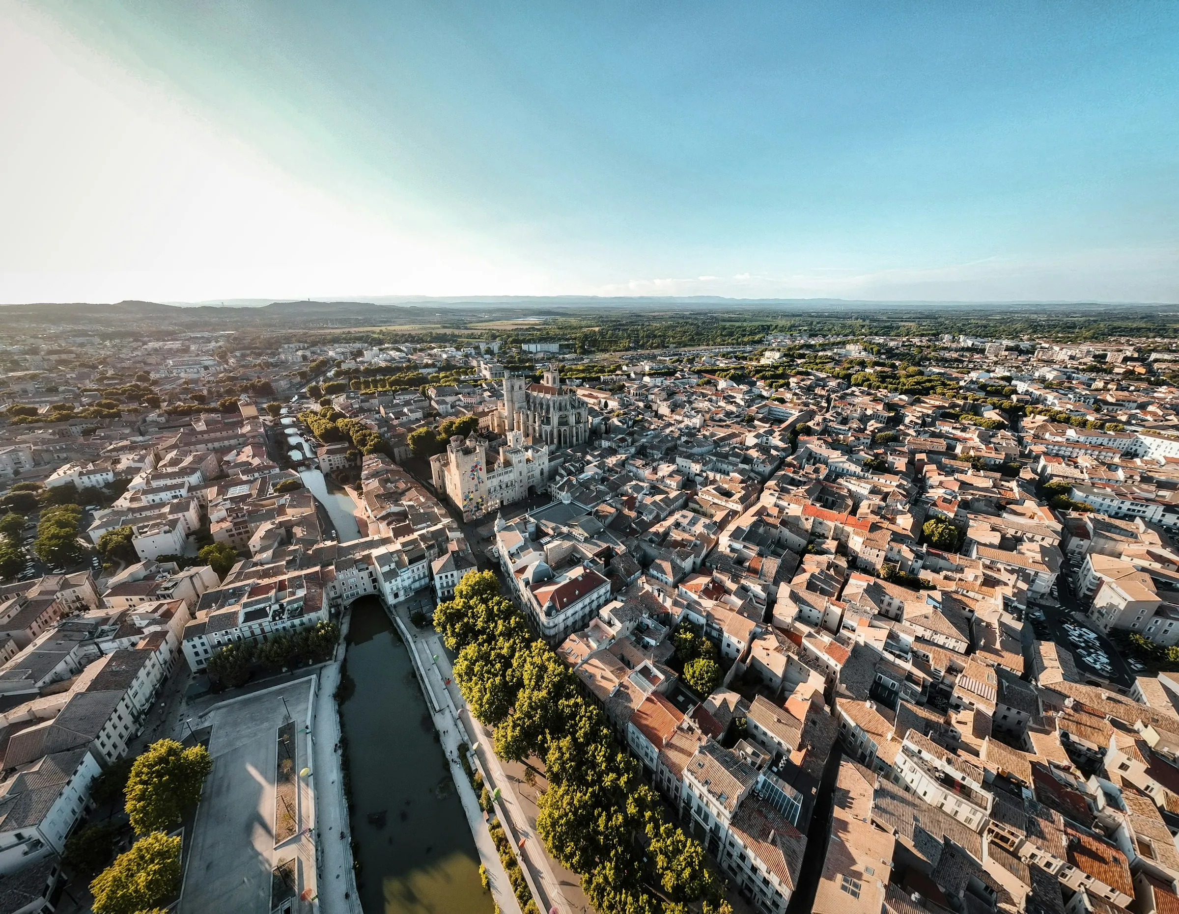 Golden-hour aerial view of Narbonne, the Canal de la Robine curving through the terracotta-roofed old town with the towering Gothic Cathedrale Saint-Just-et-Saint-Pasteur and the Palais des Archeveques at its heart, the Mediterranean plain stretching to distant hills