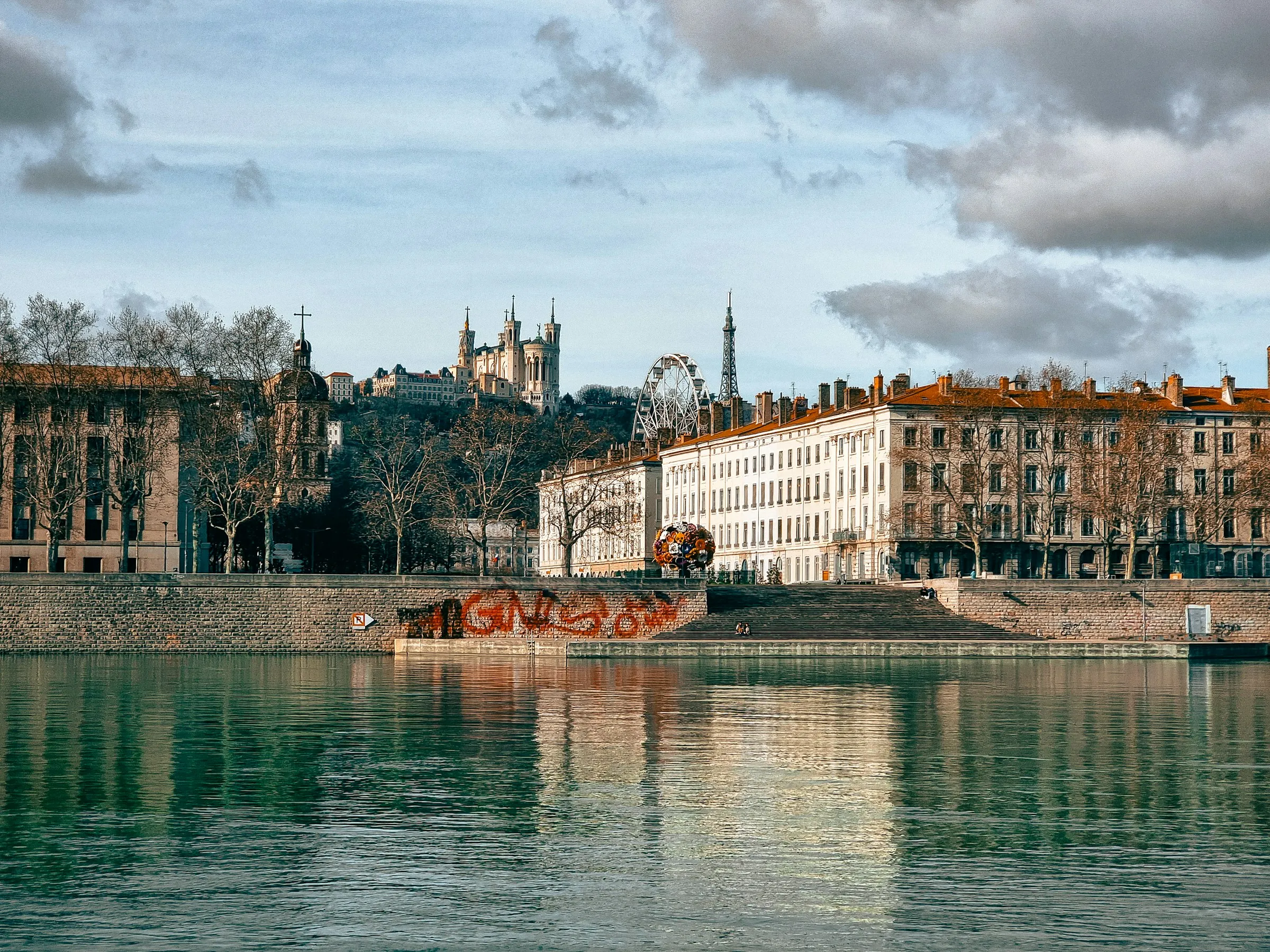 The Saone riverbank at golden hour with the white Fourviere basilica and the Tour metallique crowning the hill above the Presqu'ile's cream facades and red roofs, mirrored in the calm green water