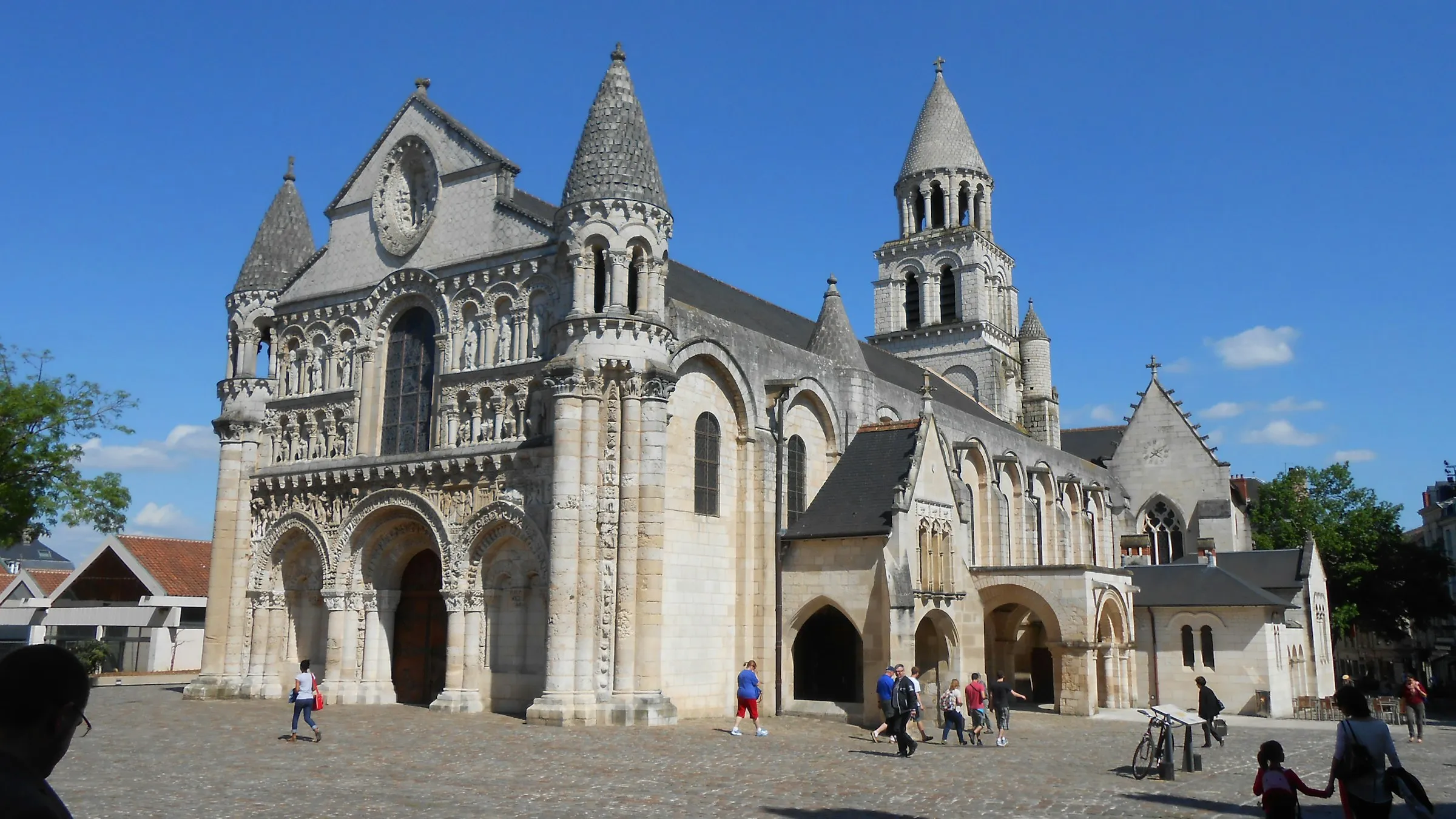 The richly sculpted Romanesque west facade and conical scaled towers of Notre-Dame-la-Grande rising above a sunlit cobbled square in central Poitiers, with people strolling past under a clear blue sky.
