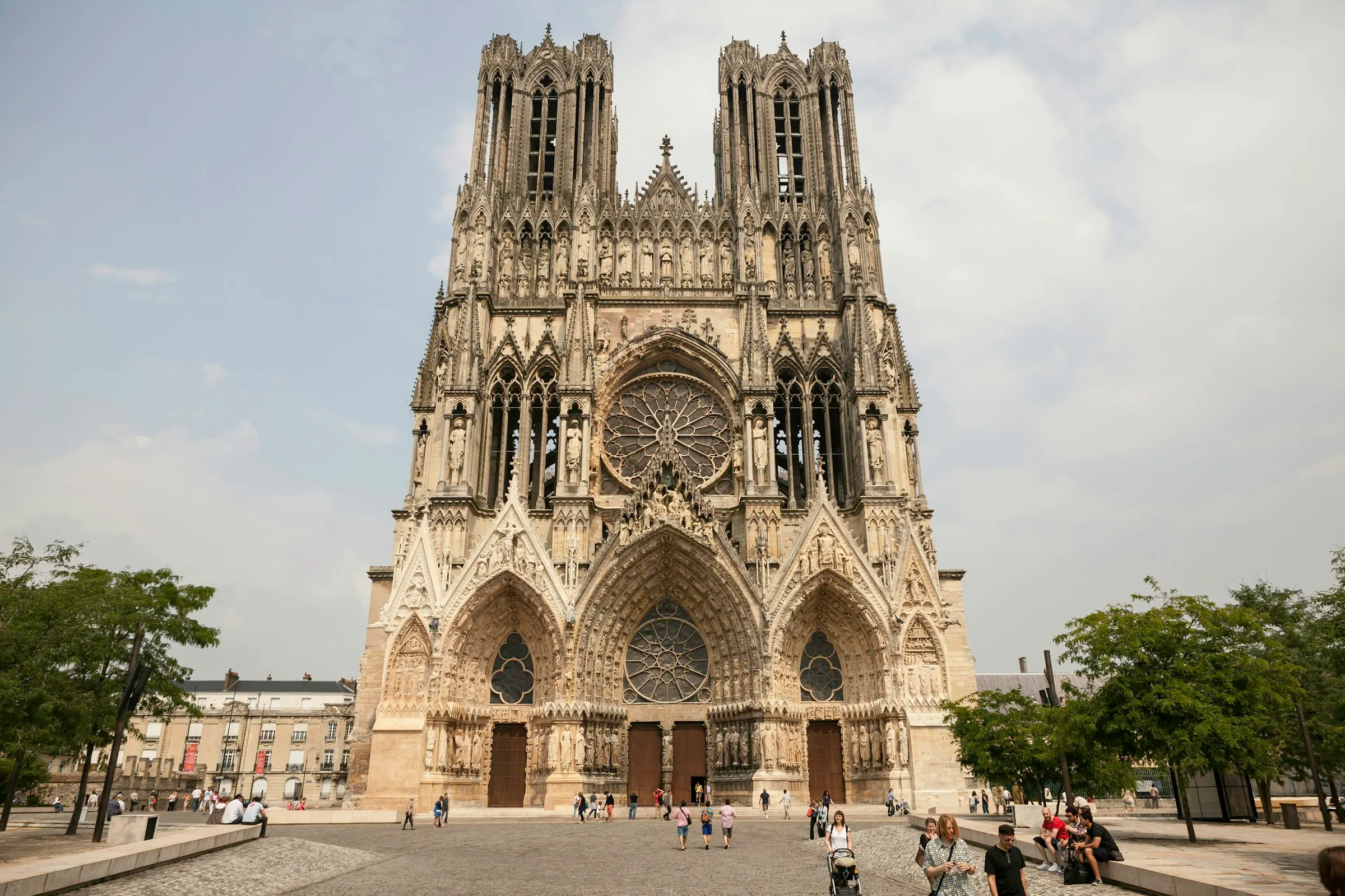 The west facade of the Gothic Notre-Dame de Reims cathedral rising over its cobbled forecourt, its twin openwork towers, gallery of kings, deeply sculpted portals and great rose window catching soft daylight as visitors cross the square below
