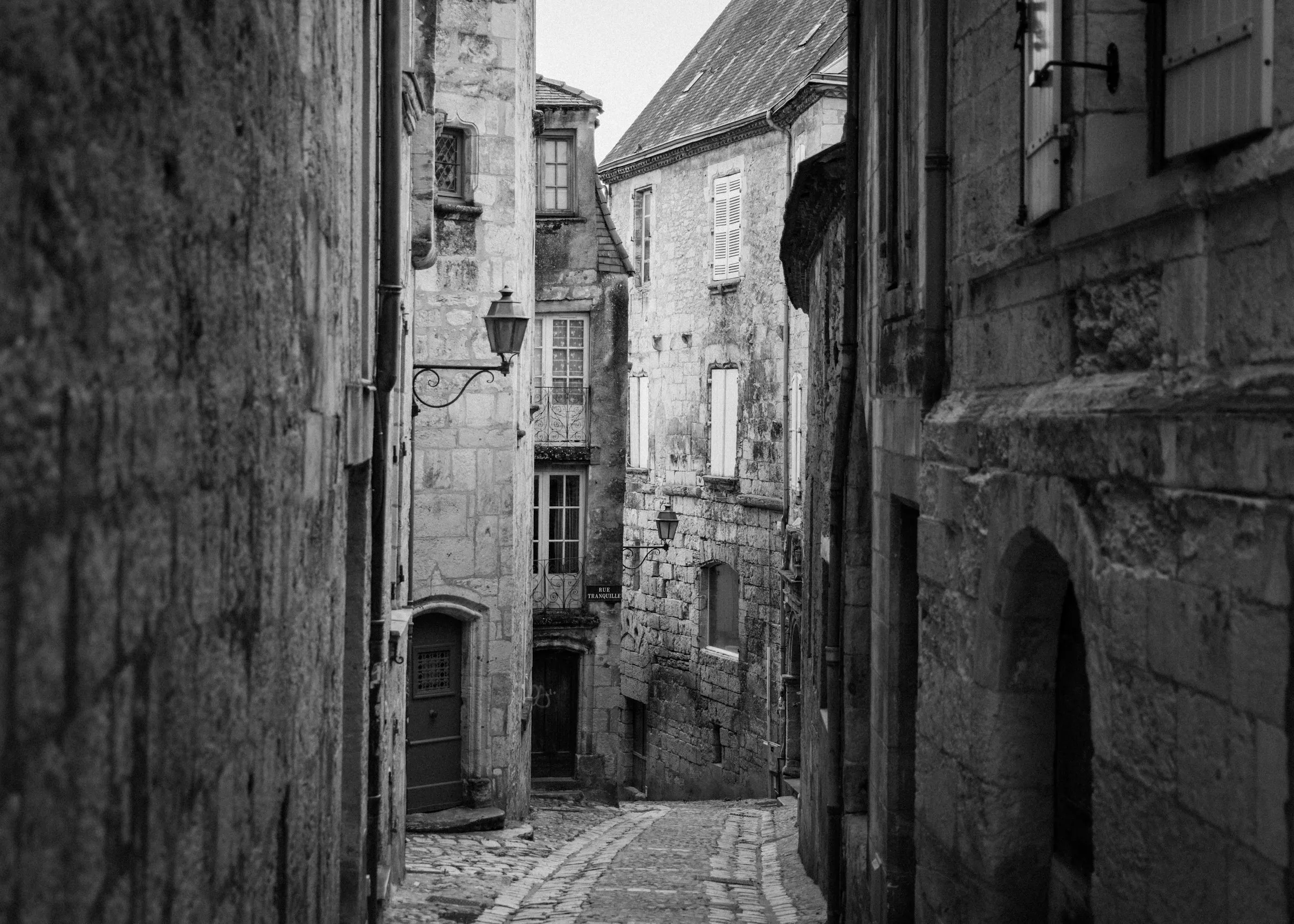 A quiet cobbled medieval lane in Perigueux's old town, lined with tall honey-stone houses and a wrought-iron street lamp, the 'Rue Tranquille' sign visible as the alley bends toward a bright opening