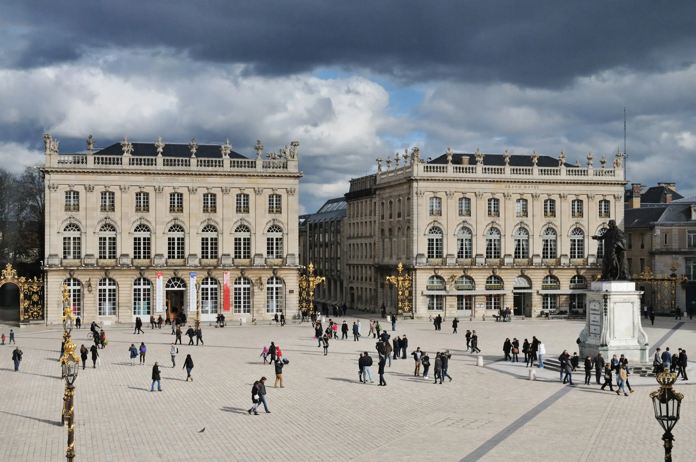 Wide elevated view across the vast cobbled expanse of Place Stanislas in Nancy, the Opera National de Lorraine and Grand Hotel flanking gilded wrought-iron gates with the Stanislas statue at right, crowds strolling under a dramatic cloud-filled sky