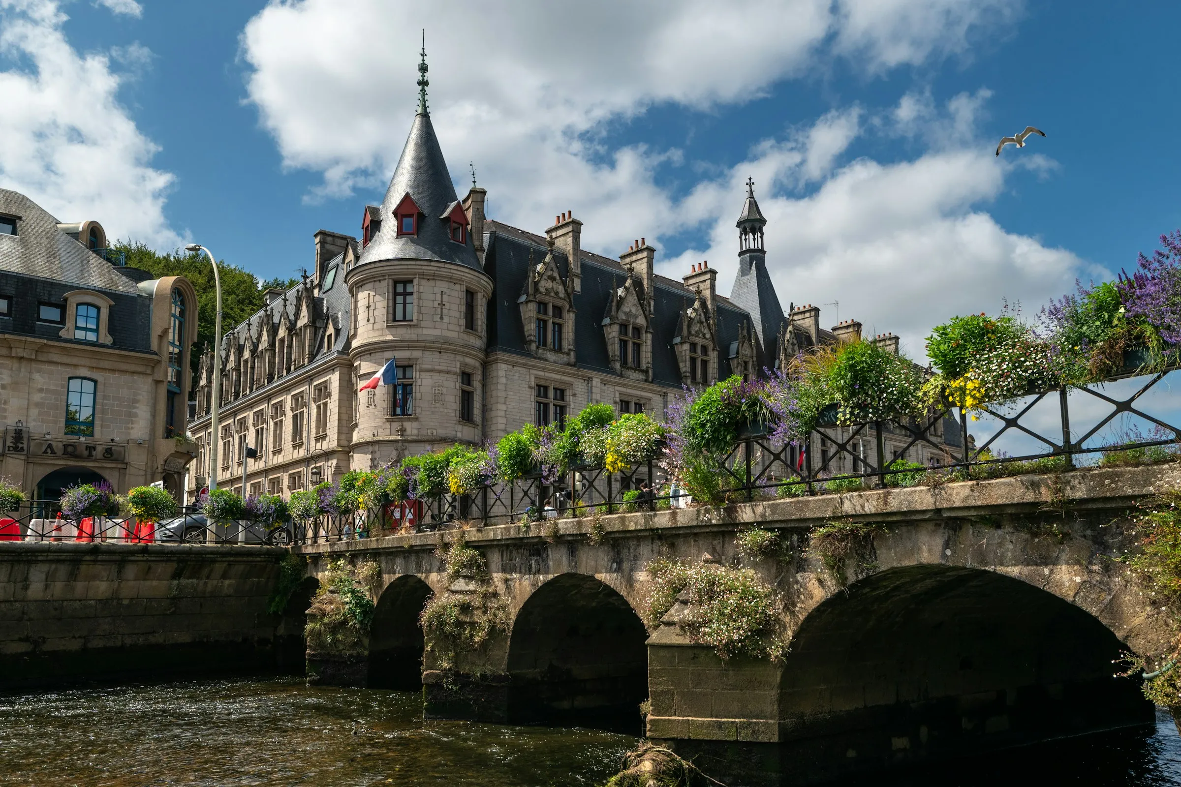 A stone turreted mansion — the Finistere prefecture — rises above the Odet river in Quimper, its low arched bridge and flower-laden iron railings reflected in the water beneath a big blue sky with a gull overhead.