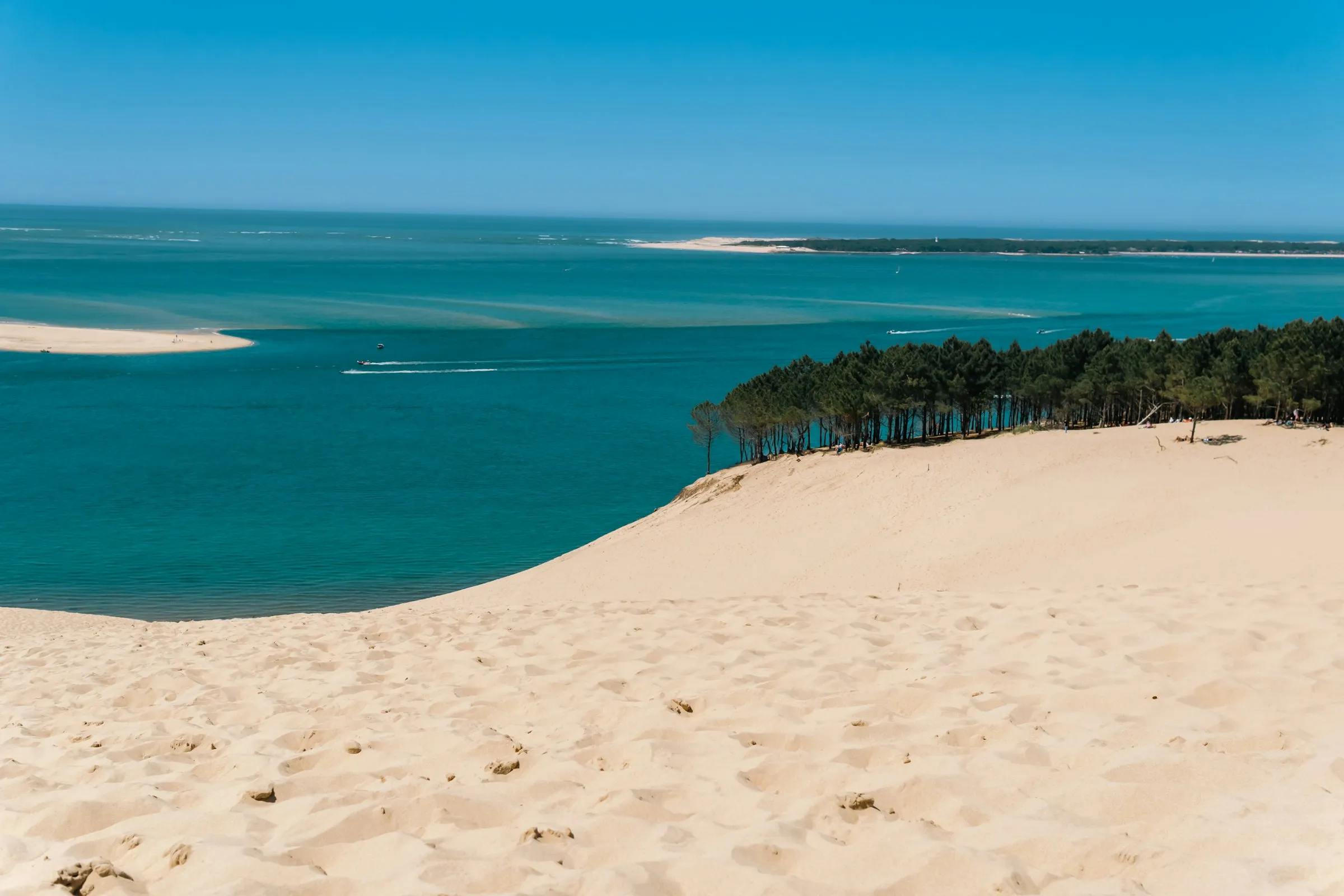 The Dune du Pilat sloping down to the turquoise Bassin d'Arcachon, a pine forest running along the sandy crest and a bright sandbank offshore under a clear blue sky.