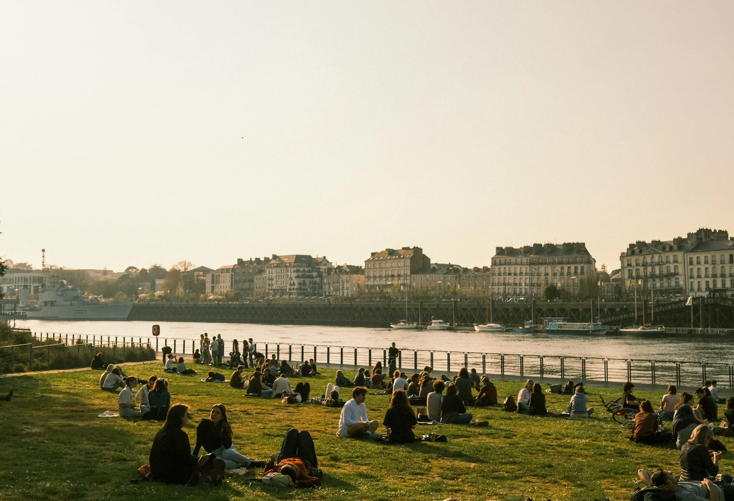 Crowds relaxing on the grassy banks of the Loire in Nantes at golden hour, with elegant stone townhouses and a moored grey naval ship lining the far quay under a soft glowing sky.