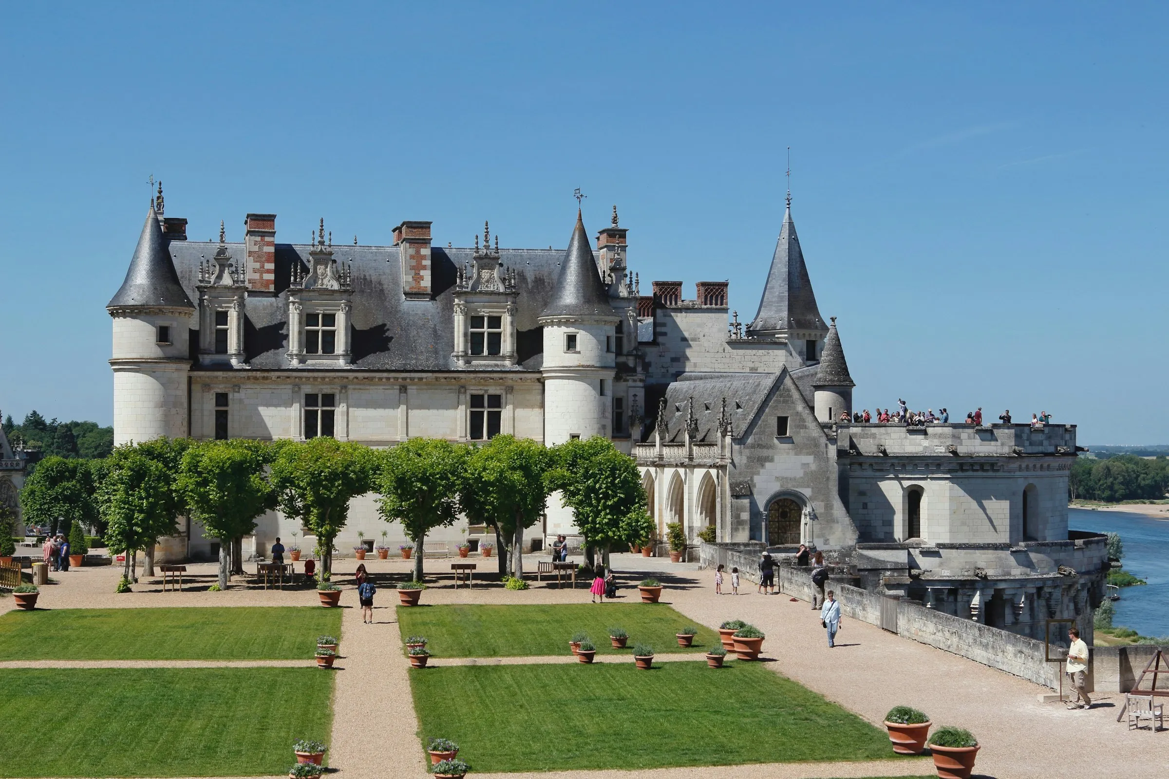 The Château Royal d'Amboise, its white-tufa Renaissance lodging and conical slate turrets rising on the ramparts high above the Loire, with the Gothic Chapelle Saint-Hubert at right and formal lawns and clipped trees on the garden terrace under a clear blue sky.