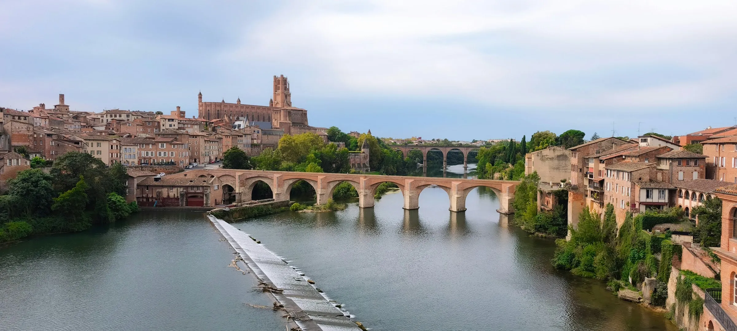 Panorama of Albi across the Tarn: the medieval red-brick Pont-Vieux and its arches spanning the calm river, with the old town's terracotta rooftops and the tower of Cathedrale Sainte-Cecile rising on the hill behind.