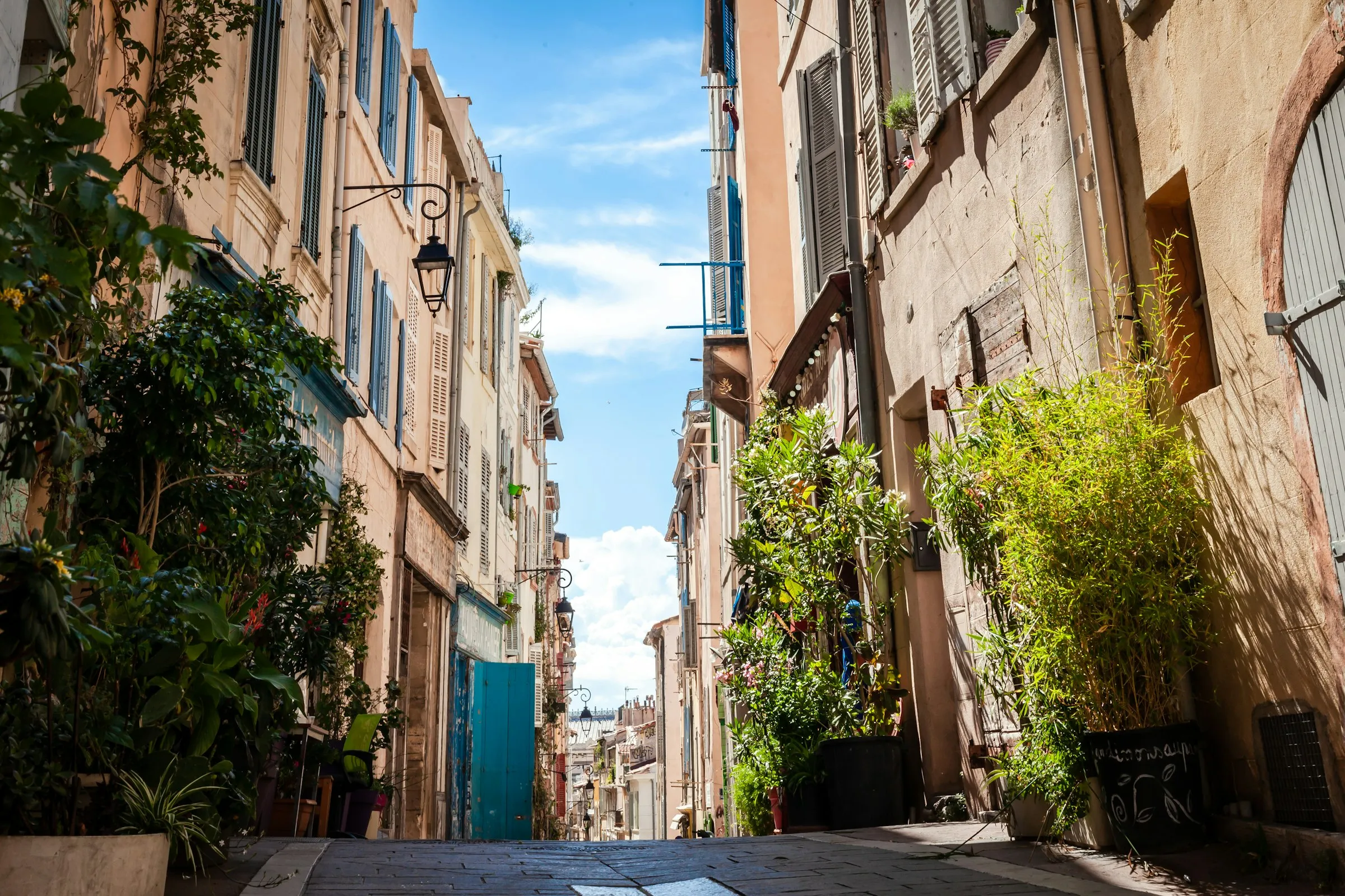 Sunlit sloping cobbled lane in Marseille's Le Panier old quarter, lined with pastel ochre and salmon townhouses, blue-painted shutters and doors, and lush potted greenery spilling out under a bright blue sky.