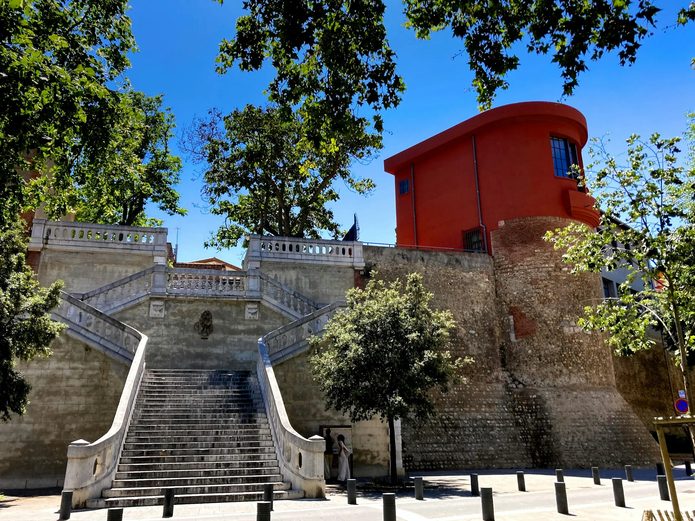 A monumental double stone staircase beside Perpignan's old bastion, built in the local banded river-pebble and red-brick masonry and topped by an ochre-red building, under a bright blue summer sky.