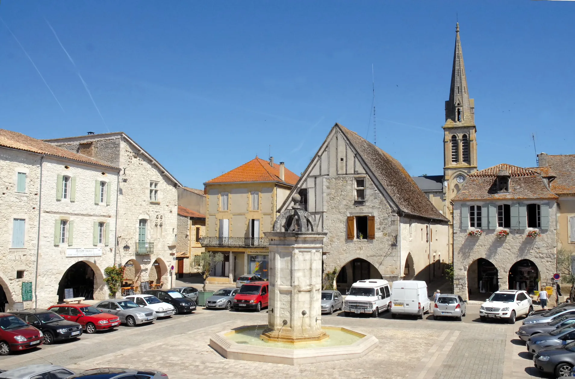 The Place Gambetta, the arcaded central square of the medieval bastide of Eymet in the Dordogne — its 17th-century stone fountain, a half-timbered house and the spire of Notre-Dame framed by the stone couverts under a bright blue sky