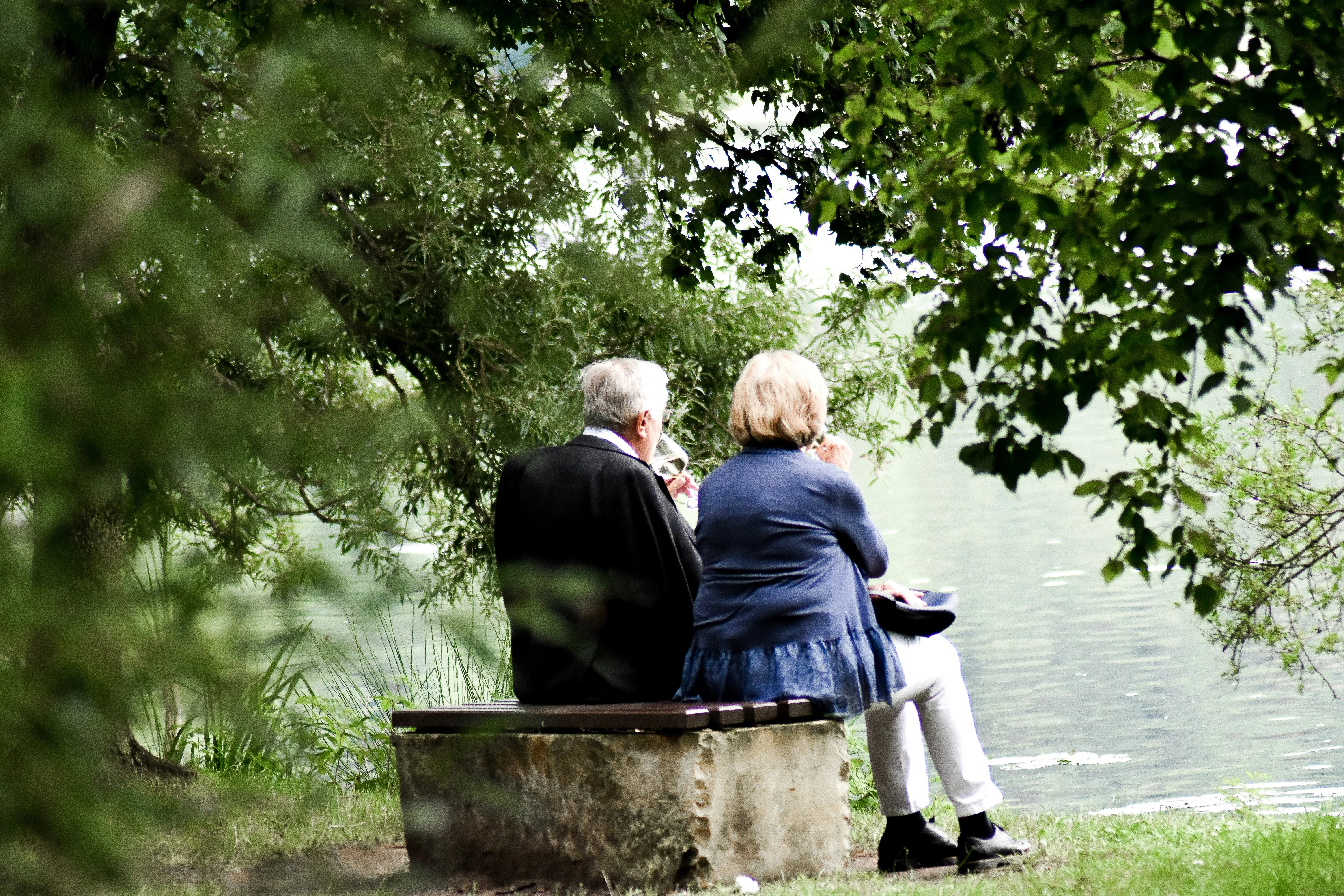An older couple sharing a glass of wine on a stone bench at the water's edge, framed by trees